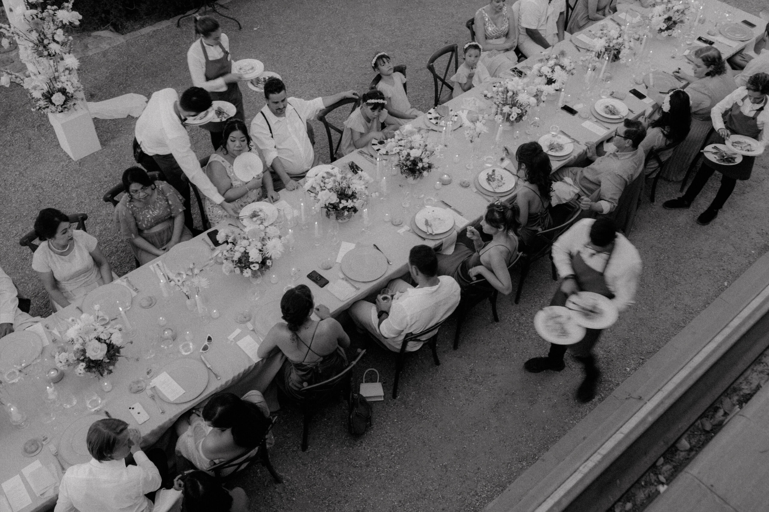 Vogelperspektive auf ein elegantes Hochzeitsdinner im Freien in Düsseldorf, mit Gästen in festlicher Kleidung, die um eine lange Tafel mit weißer Tischdecke und Blumendekoration sitzen, eingefangen in monochromer Fotografie für ein klassisches Flair.