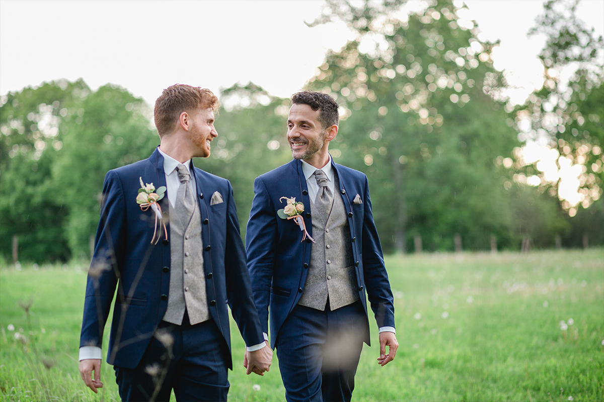 Hochzeitspaar mit zwei Bräutigamen geht über eine Wiese, halten Hände und lächeln sich an