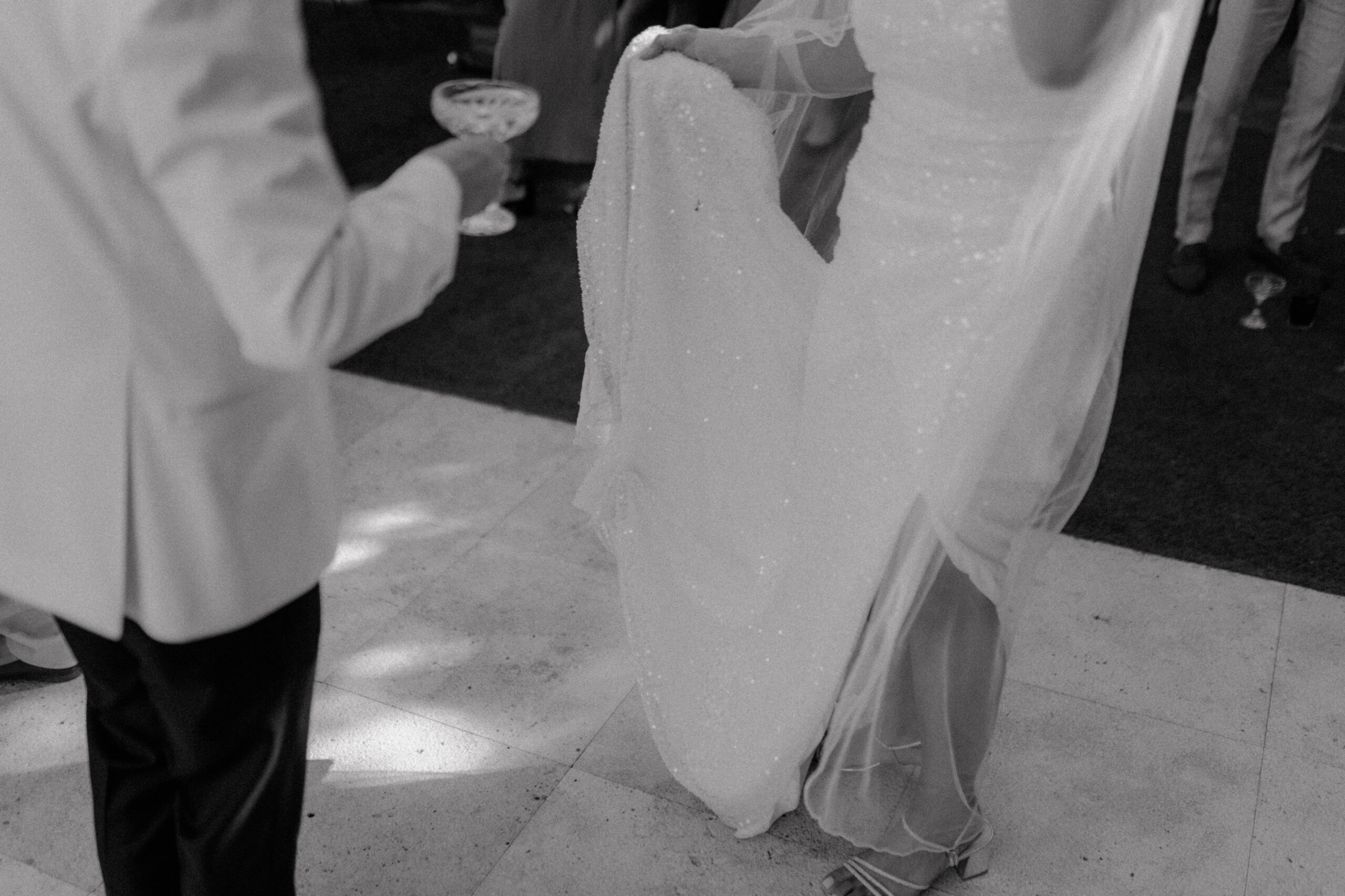 Detailaufnahme der Umarmung eines Brautpaares auf ihrer Hochzeitsfeier in Köln, fokussiert auf die glitzernde Brautkleid-Schleppe und die elegante Handhaltung, in Schwarz-Weiß für eine zeitlose Ästhetik.