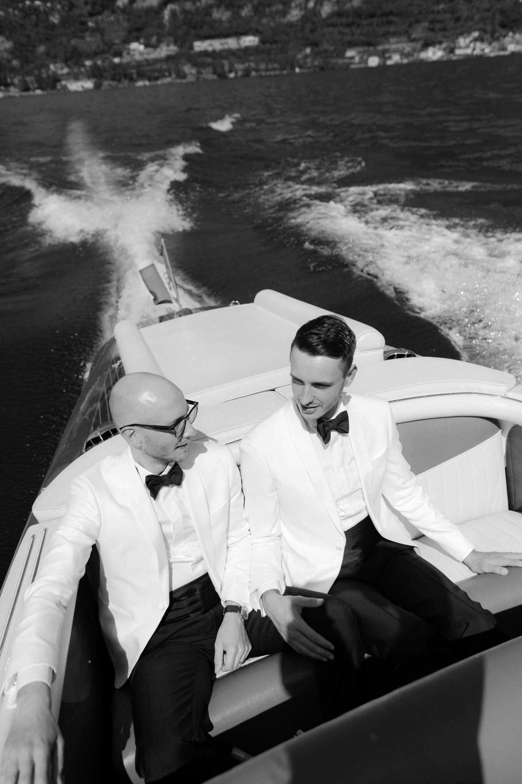 Elegantes Hochzeitspaar in weißen Smokings fährt in einem Boot über den See, stilvoller Moment nach der Hochzeitszeremonie.