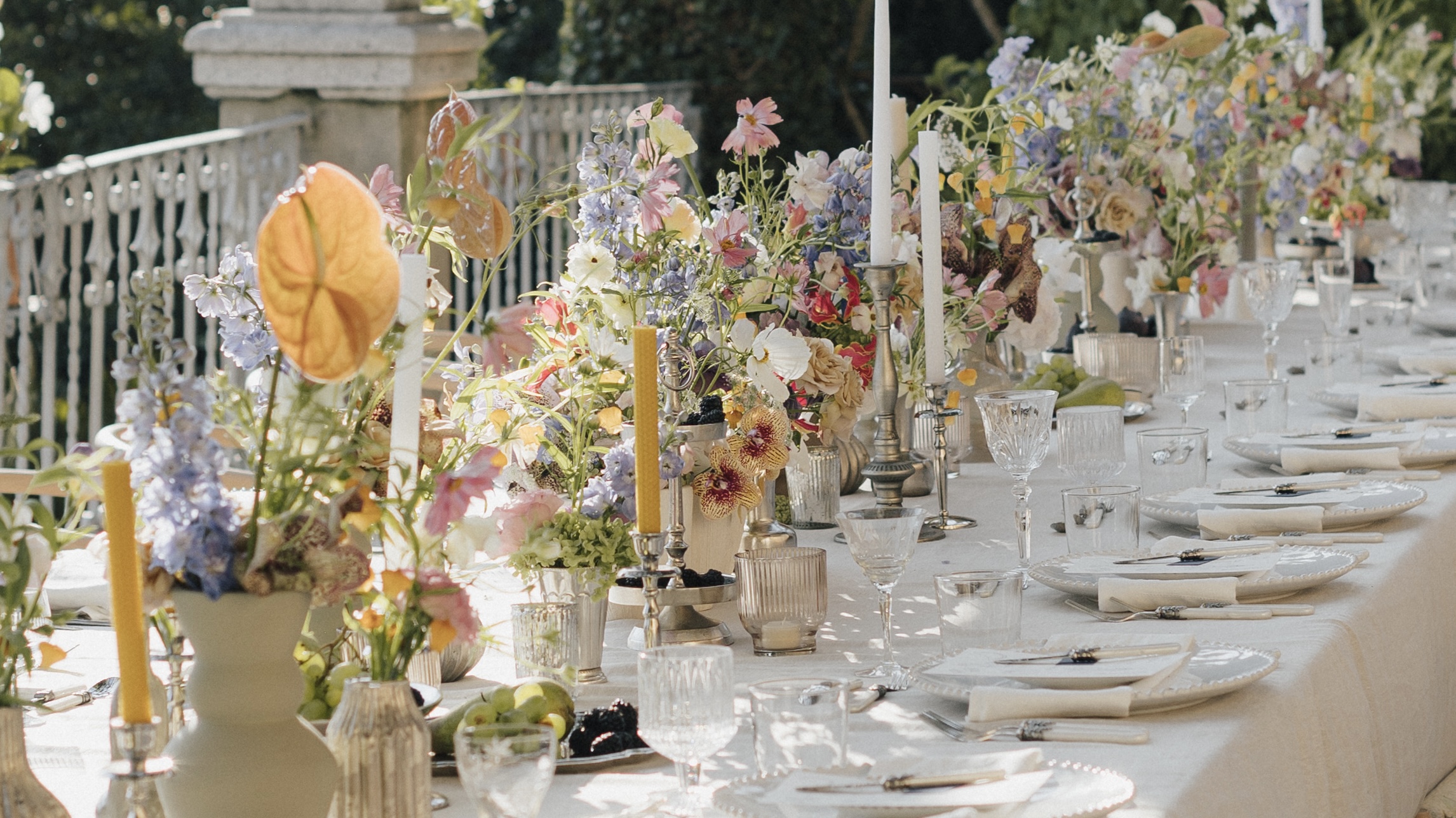 Hochzeitstafel im Freien mit reichhaltiger Blumendekoration, Kerzen und feinem Geschirr.