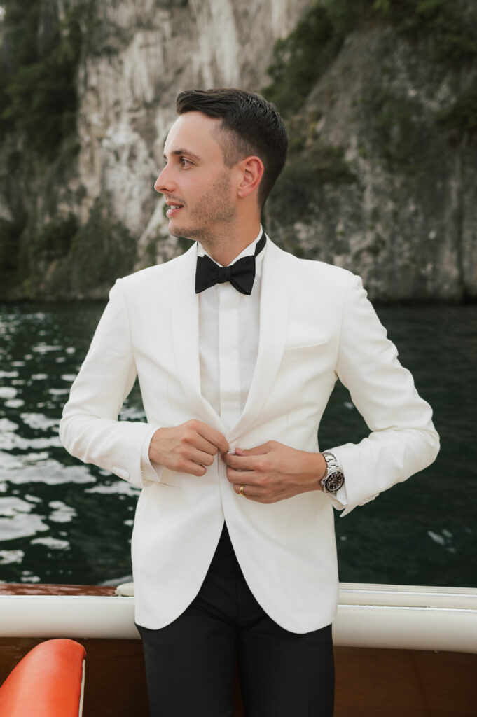 Eleganter Bräutigam in weißem Smoking mit Fliege auf einem Holzboot, vor malerischer Felskulisse in Italien – stilvolles Hochzeitsfoto am Wasser. Destination Wedding.