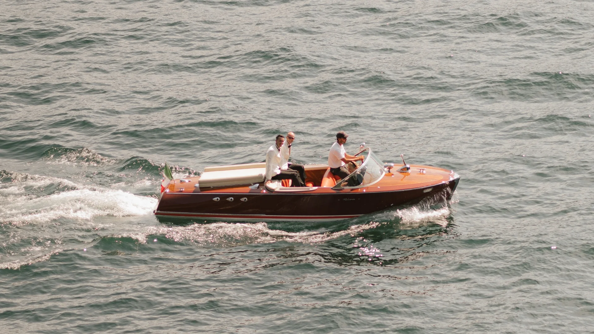 Brautpaar in eleganter Kleidung auf klassischem Holzboot bei Bootsfahrt in Italien, umgeben von sanften Wellen – stilvolle Hochzeitsmomente am Wasser